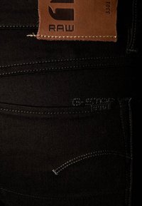 Musta farkkuhousut, joissa on nahkainen merkki vyötäröllä. Yksityiskohtainen ompelu ja hillitty kohokuvioitu logo korostavat designia.