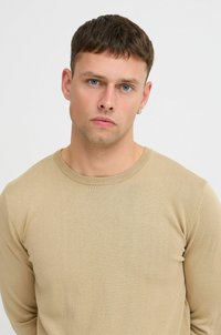 Suéter de punto beige con cuello redondo y mangas largas, que presenta una textura suave y un diseño ajustado. Sin patrones ni acentos visibles.