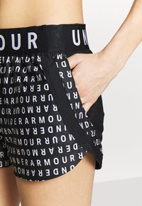 Shorts de sport noirs avec le texte "UNDER ARMOUR" en blanc répété, taille élastique, poches latérales, portés par une personne ayant la main à l'intérieur de la poche.