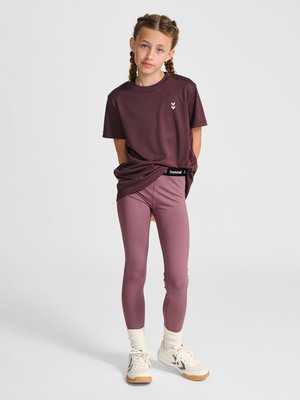 Ung pige med flettet hår iført en mørkerød t-shirt og mauve leggings, stående med hænderne bag ryggen mod en ensfarvet baggrund.