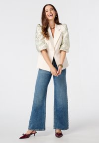 Lichtbeige blazer met gepofte, gemusterde mouwen, gecombineerd met wijd uitlopende blauwe jeans en kastanjegekleurde muiltjes, wat een casual, moderne uitstraling geeft.