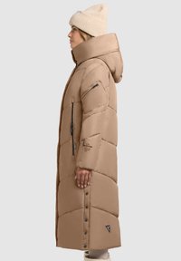 Hosszú, bolyhos bézs kabát kapucnival, oldalsó cipzárakkal és elöl patentgombokkal, texturált anyagból készült, a ujján logóval.