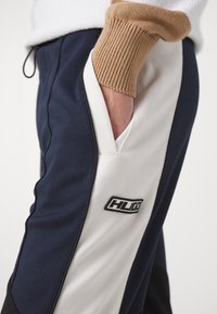 Marinblå och vita träningsbyxor med en stilren design, försedda med en sidficka och en svart logotyp. Texturerad stickad mudd i ljusbrunt.