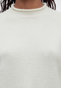 Gros plan d'une personne portant un pull en tricot côtelé blanc avec un col rond et des cheveux tressés visibles sur les épaules.