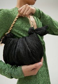 Bolso negro acolchado texturizado con un asa de cadena de oro trenzada, que presenta un acento anudado en un lado. Llevado contra un vestido verde.