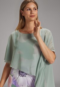 Donna con blusa trasparente verde chiaro e gonna floreale che alza una mano vicino al viso, guardando leggermente verso l'alto contro uno sfondo grigio uniforme.