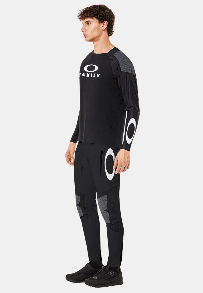 Homme debout de profil portant des vêtements de sport noirs Oakley avec de grands logos blancs sur la poitrine, la manche et le pantalon, sur un fond uni.