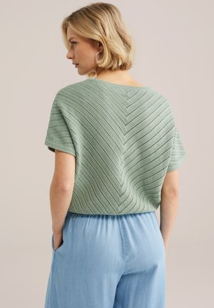 Blonde sieviete, kas valkā sudraba riņķa auskarus, zaļu īsroku trikotāžas topu ar zivtiņrindu rakstu un gaiši zilas augstas jostas bikses, ar rokām kabatās.