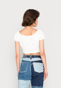 Top branco curto com mangas bufantes, feito de tecido leve. Combinado com calções de ganga em patchwork com bordas desfiadas e múltiplos bolsos.