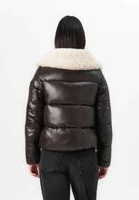 Vrouw in een donkerbruine pufferjack met een grote crèmekleurige sherpa-kraag, gecombineerd met zwarte denim jeans, met de rug naar de kijker gekeerd.