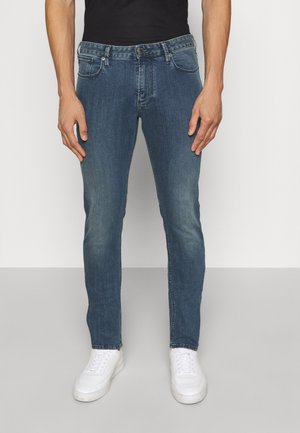 Homme portant un jean slim bleu, des baskets blanches et une chemise noire, debout devant un fond clair uni.