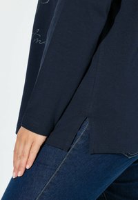 Maglione blu navy a maniche lunghe con fessure laterali, scollo rotondo e texture di cotone morbido, abbinato a jeans blu scuro.