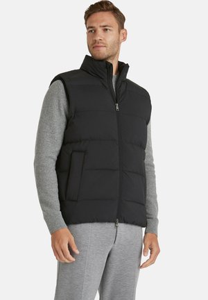 Man draagt een zwart donsjack over een lichtgrijze trui en broek, staat met ontspannen handen en kijkt zijwaarts tegen een witte achtergrond.