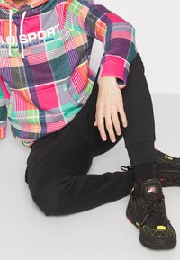 Hoodie a quadri colorati con toni di rosa, verde e viola, dotato di tasca a marsupio e grafiche bianche. Pantaloni neri aderenti e sneakers nere.