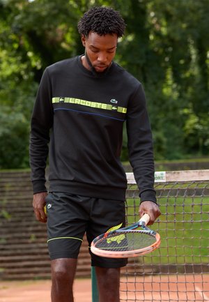 Un sweat-shirt noir avec une bande graphique verte et bleue, associé à un short noir avec une accentuation verte. Tenant une raquette de tennis près d'un filet.