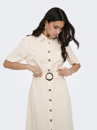 Kvinne som justerer belte på beige knappet kjole med korte ermer, rund skilpaddeskallspenne og matchende øredobber mot ensfarget bakgrunn.