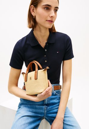Jeune femme portant un polo noir et un jean bleu, tenant un petit sac tissé beige avec des poignées en cuir marron.