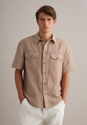 Junger Mann, der ein beiges kurzärmliges Hemd mit Brusttaschen und weiße Hose trägt, steht mit den Händen in den Taschen.