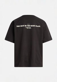 Zwart katoenen T-shirt met korte mouwen en een ronde hals. Tekst op de achterkant luidt "DIT MOET DE JUISTE PLEK ZIJN" in het wit.