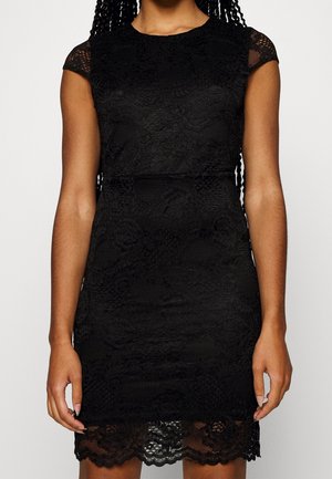 Sort tætsiddende blondekjole med kappelignende ærmer og bølget nederkant båret af person med langt flet hår.