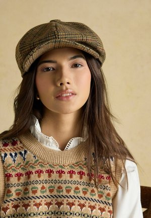 Braune kariertes Mütze mit gebogenem Schirm. Beiger Pullover mit bunten Mustern von Bäumen, Blumen und Tieren, darüber ein weißes Hemd mit Kragen.