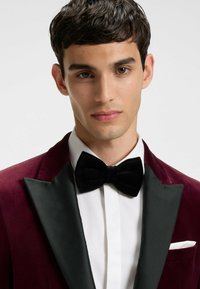 Bordeauxrotes Samt-Tuxedojacket mit schwarzen Satinrevers, weißem Hemd und schwarzer Samtfliege. Mit einer Brusttasche und taillierter Passform.