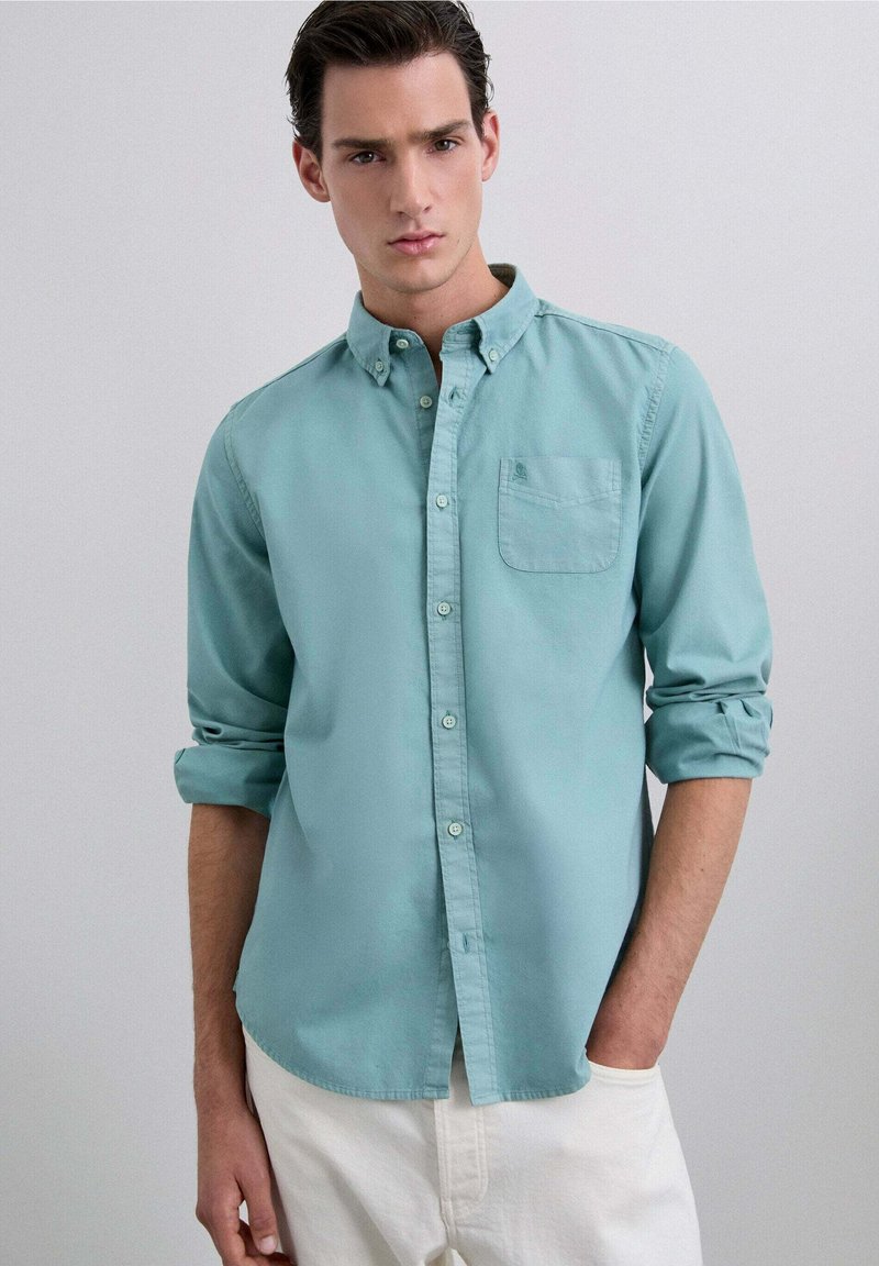 Camisa abotonada de color turquesa claro, hecha de algodón, con un bolsillo en el pecho y mangas enrolladas, combinada con pantalones blancos.