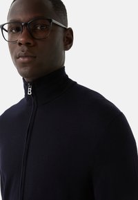 Mężczyzna w czarnych okularach i ciemnoniebieskim swetrze zapinanym na zamek, z wysokim kołnierzem, na jednolitym jasnym tle.