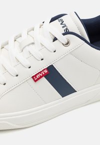 Baskets blanches Levi's avec une bande et une doublure bleu marine, lacets blancs, étiquette rouge avec logo Levi's sur le côté et œillets métalliques.
