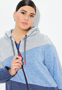 Mikina s kapucí a na zip v šedé, světle modré a navy barvě. Obsahuje šňůrky s červenými akcenty a texturovanou tkaninu.