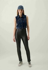 Colete de ganga sem mangas, lavagem escura, com bolsos frontais; combinado com calças de couro pretas justas; sapatilhas cinzentas; boné azul.