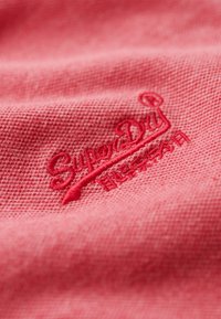 Tela roja con un tejido texturizado, con un logotipo bordado de "SuperDry" en hilo rojo a juego cerca del centro.