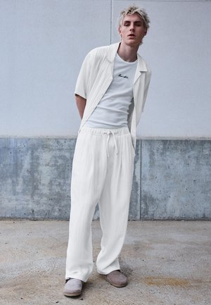 Jonge persoon met lichtblond haar, gekleed in losse witte broek, een wit overhemd met korte mouwen over een wit hemdje, en grijze schoenen, staand.