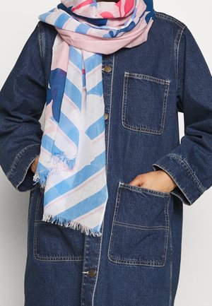 Personne avec les mains dans les poches d'une veste en denim foncé, portant une écharpe blanche, rose et bleue à motifs, nouée de façon lâche autour du cou.