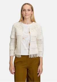 Gilet en crochet crème au design ajouré, avec des manches trois-quarts et des détails en frange, assorti d'un t-shirt floral blanc et d'un pantalon marron.