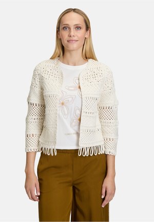 Gilet en crochet crème au design ajouré, avec des manches trois-quarts et des détails en frange, assorti d'un t-shirt floral blanc et d'un pantalon marron.