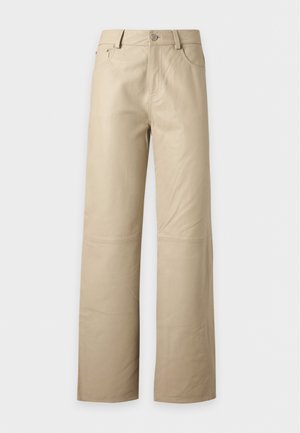 Kvinde iført beige jakke og vide bukser, lyseblå skjorte, sort taske på skulderen og sorte sko, stående mod en ensfarvet baggrund.