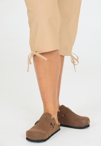 Ruske suède klompe med en blød struktureret finish, der har en lukket tå og et dekorativt bånd. Sæt sammen med lysebrune beskårne bukser, der er bundet i kanten.