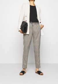 Beige sako, černý top, kostkované kalhoty se bočními pruhy a černé sandály. Výrazná černá kabelka s texturovaným povrchem.