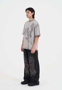 Camiseta gris de gran tamaño con gráficos de águila y texto. Vaqueros negros con parches de patrones intrincados. Calzado negro con superficie texturizada.