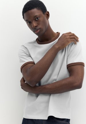 Young man with short curly hair wearing a white t-shirt with brown trim, standing with arms crossed and one hand on his shoulder.