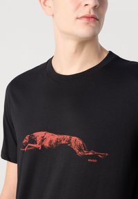 Svart bomulls T-skjorte med grafikk av en hoppende hund i rødt; korte ermer, rund hals og "HUGO"-logo under designet.
