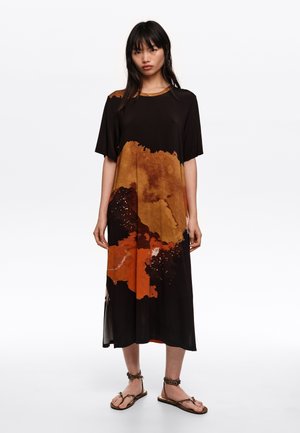 Kobieta stojąca w czarnej sukience midi z brązowo-pomarańczowymi abstrakcyjnymi wzorami, ubrana w brązowe sandały z wiązaniem wokół kostki na jednolitym białym tle.