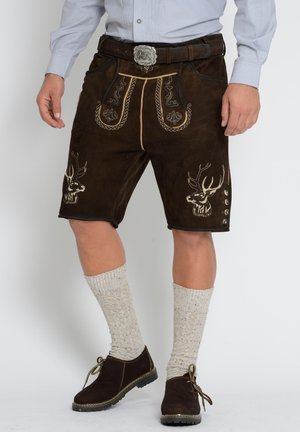 Mężczyzna noszący tradycyjne brązowe skórzane lederhosen z haftowanymi głowami jeleni, szare skarpety do kolan, ciemne buty i jasnoniebieską koszulę.
