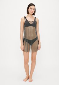 Copertura in rete nera trasparente con design senza maniche, caratterizzata da un motivo a pois e una vestibilità ampia, indossata sopra un perizoma nero.