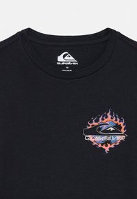 T-shirt preta de algodão com gola redonda. Apresenta um logotipo gráfico multicolorido rodeado por chamas laranjas e azuis no lado esquerdo do peito.