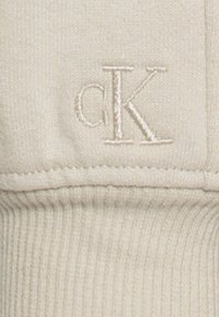 Stickad bomullströja i krämfärg med ribbad struktur. cK-logotypen är broderad i en något mörkare nyans och placerad på bröstet.