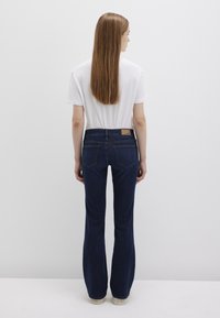 Dark blue flared jeans with a mid-rise waist, featuring a subtle brand label on the back pocket and a smooth denim texture. Paired with a white t-shirt.