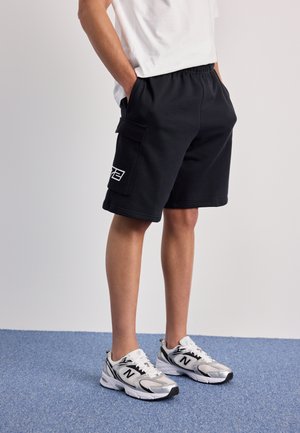 Person iført sorte cargoshorts med klaplomme og hvide New Balance sneakers, stående på blåt tæppe.
