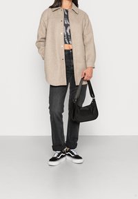 Donna che indossa un cappotto beige con bottoni, un crop top nero a fantasia, jeans neri a risvolto, sneaker bianco e nere, mentre tiene una borsa nera.